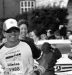 A male athlete in race gear smiles as he runs toward the finish line, wearing sunglasses, a cap, and bib 1986. Spectators on the left cheer and clap. The event feels energetic with banners, a parked car, and even a villa mia review sign in the background. {{brizy_dc_image_alt imageSrc=