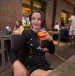 A woman with long dark hair, wearing a black dress with white polka dots, sits at an outdoor restaurant table holding an orange cocktail. She is smiling slightly and looking to her left. The background shows other diners and warm lighting. {{brizy_dc_image_alt imageSrc=